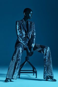 a woman sitting on top of a stool wearing silver sequin pants and a jacket