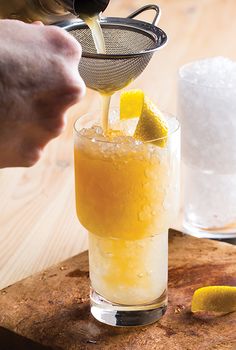 a person pours a drink into a glass with ice and lemon wedges on the side