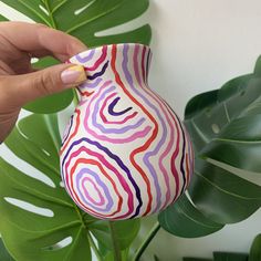 a hand holding a colorful vase next to a plant