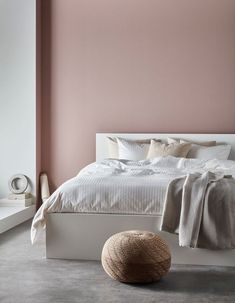 a white bed sitting next to a pink wall and a round ottoman on the floor