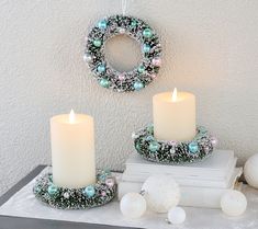 two white candles are sitting on top of some books and ornaments in front of a wreath