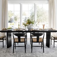 a dining room table with chairs and plates on it, in front of large windows