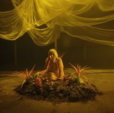 a woman sitting on top of a pile of dirt next to plants and flowers in front of a curtain