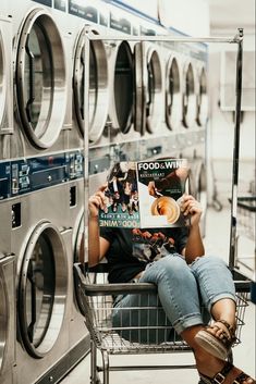 a woman is sitting in a shopping cart reading a magazine next to washing machine doors