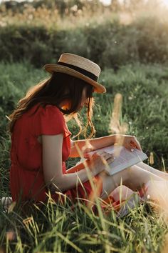 a woman sitting in the grass reading a book and looking at her phone while wearing a straw hat