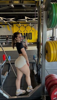 a woman standing in front of a barbell rack holding a water bottle and looking at her cell phone