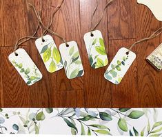 four tags are hanging from twine on a wooden floor with leaves and branches painted on them