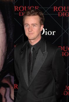 a man in a black suit and tie at a red carpeted room with a movie poster behind him