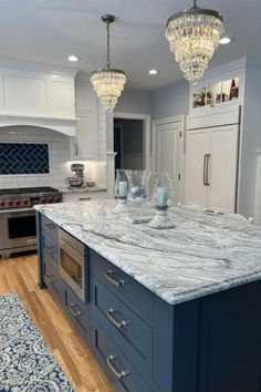 a large kitchen island with marble counter tops and blue cabinets in the center is flanked by two crystal chandeliers