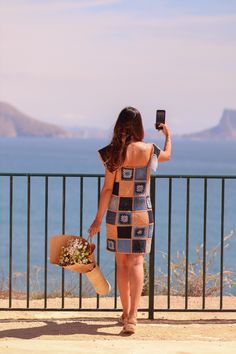 a woman taking a photo with her cell phone while standing next to a fence and looking at the ocean