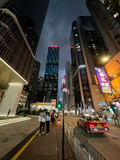 people are walking on the sidewalk in front of tall buildings at night time, with cars driving down the street