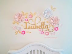 a white crib with pink and gold flowers on the wall, next to a baby's name sign