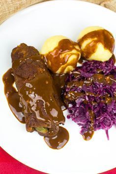 a white plate topped with meat covered in gravy next to purple cabbage and potatoes