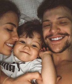 a man and woman smile as they lie in bed with a baby on their lap