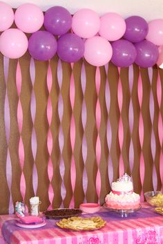 a table topped with cake and balloons next to a wall covered in streamers filled with pink and purple balloons