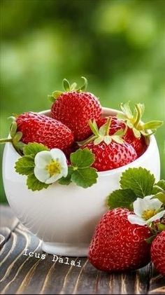 strawberries in a white bowl on a wooden table with green leaves and flowers around them