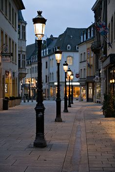 an empty street at dusk with several lamps on the sidewalk and buildings in the background