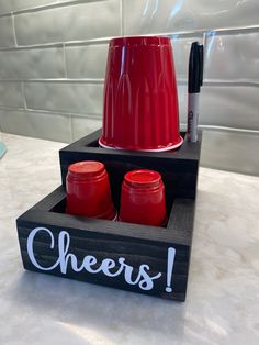 there is a red cup and two jars in a holder on the counter top with writing cheers