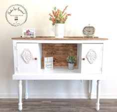 a white cabinet with two drawers and some plants on top, next to a clock