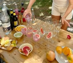 a woman is pouring wine into glasses on a wooden table with fruit and bottles in the background