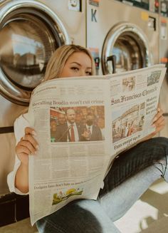 a woman sitting in front of a dryer while reading a newspaper with her feet on the floor