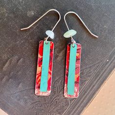 two rectangular shaped earrings with blue and red designs on them sitting on a piece of fabric