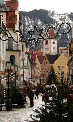 people are walking down the cobblestone street in front of some buildings and christmas trees