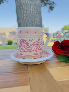 a pink and white coffee cup sitting on top of a wooden table next to a red rose