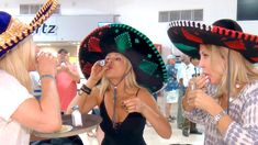 three women wearing sombreros and eating food at an event in a building with other people standing around