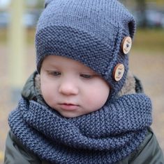 a little boy wearing a knitted hat and scarf with buttons on his head, standing in a park