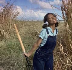 a woman standing in a field holding a baseball bat and wearing overalls with her eyes closed