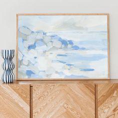 an abstract painting is displayed on top of a wooden cabinet next to a blue vase