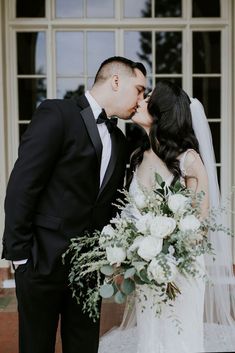 a bride and groom kissing in front of a building with white flowers on the bouquet