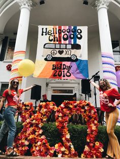 two women are standing in front of a building with flowers and balloons on the lawn