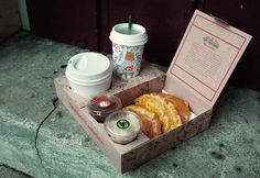 an open box with some food and drinks on the ground next to a cup of coffee