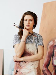 a woman holding two paintbrushes in her hand while standing next to a painting