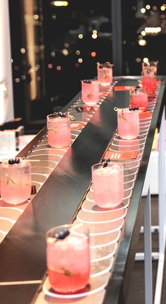 several pink drinks lined up on a metal bar with lights in the backround