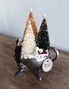 two small christmas trees in a silver cup on a table with a tag hanging from it