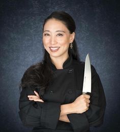 a woman holding a large knife in her right hand and smiling at the camera, against a dark background