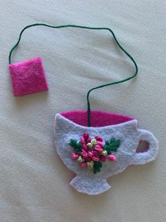 a felt tea cup with pink flowers and green leaves is hanging from a string on a white surface