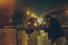 two people standing next to each other on a sidewalk at night with street lights in the background