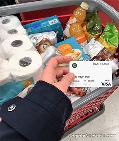 a person holding a credit card in front of a shopping basket filled with food and toilet paper