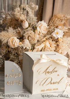 a white box with gold lettering on it next to a bouquet of flowers and cards