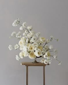 an arrangement of white flowers in a vase on a wooden stand against a gray wall