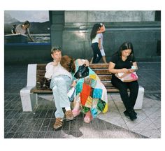 three people sitting on a bench in the middle of a city square, one holding a purse