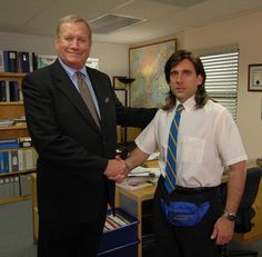 two men shaking hands in an office with bookshelves and desks behind them