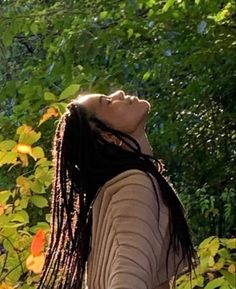 a woman with dreadlocks standing in front of some trees and bushes looking up at the sky