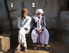 two old men sitting on a bench in front of a building with a wooden stick