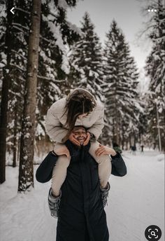 a woman carrying a child on her shoulders while standing in the snow near some trees