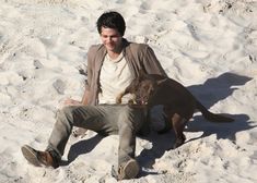 a man sitting in the sand with a dog on his lap next to him,